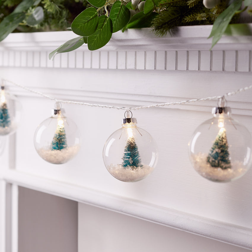 An einem Kamin aufgespannte Lichterkette mit Weihnachtsbaumkugeln aus Glas mit jeweils einem Mini-Weihnachtsbaum und Kunstschnee im Inneren