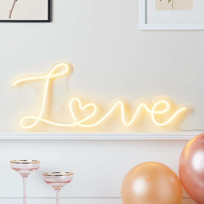 Warmweiß leuchtender Neonschriftzug "Love" an einer weißen Wand, davor rosa Champagnergläser und Luftballons
