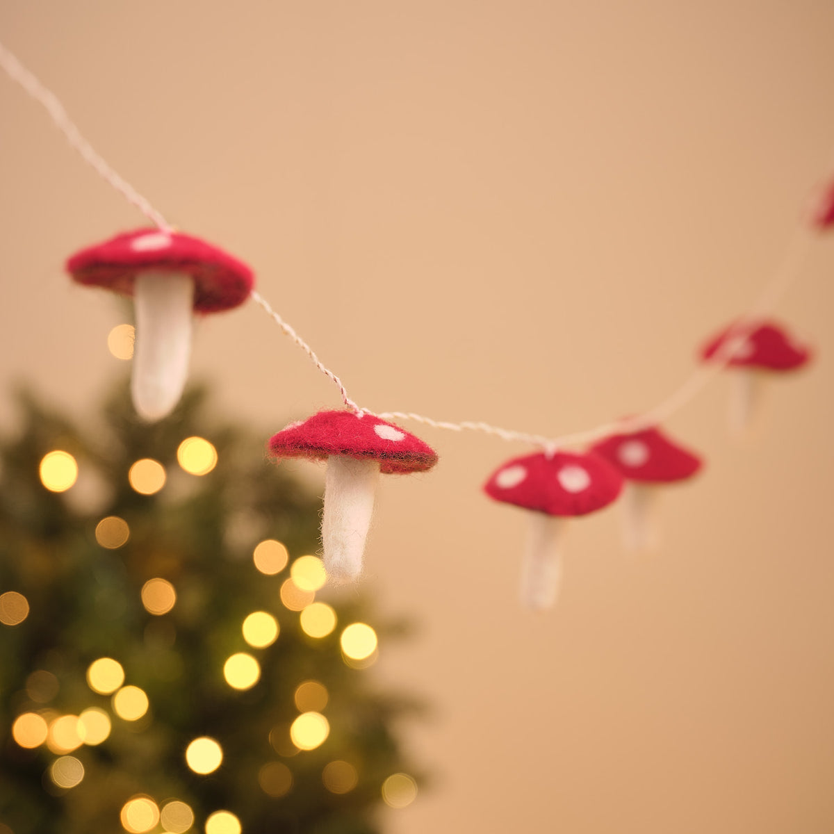 Fliegenpilz-Lichterkette aus Filz mit mehreren rot-weißen Pilzen, im Hintergrund unscharf ein beleuchteter Weihnachtsbaum.