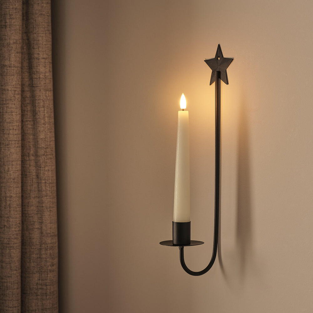 Wandmontierter LED-Kerzenhalter mit Metallstern-Aufsatz und cremefarbener LED-Kerze, schwarzes Metallgestänge, an grauer Wand montiert.
