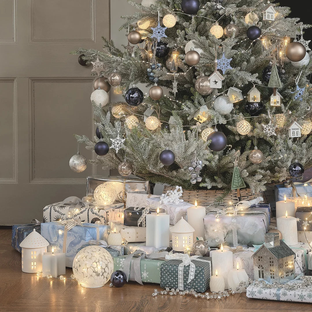 Großer geschmückter Weihnachtsbaum mit grauem Tannengrün, beige, silberne und dunkelblau Kugeln, blauen Schneeflocken, beleuchteten Häuschen-Ornamenten, darunter verpackte Weihnachtsgeschenke, weiße LED-Kerzen und Dekoration.