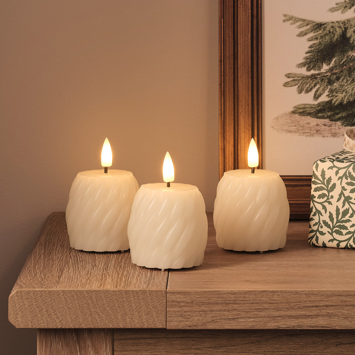 Drei weiße LED-Kerzen mit wellenförmiger Textur und warmweißen LED-Flammen auf Holztisch, beige Leinenserviette, verschwommener Tannendekorations im Hintergrund.