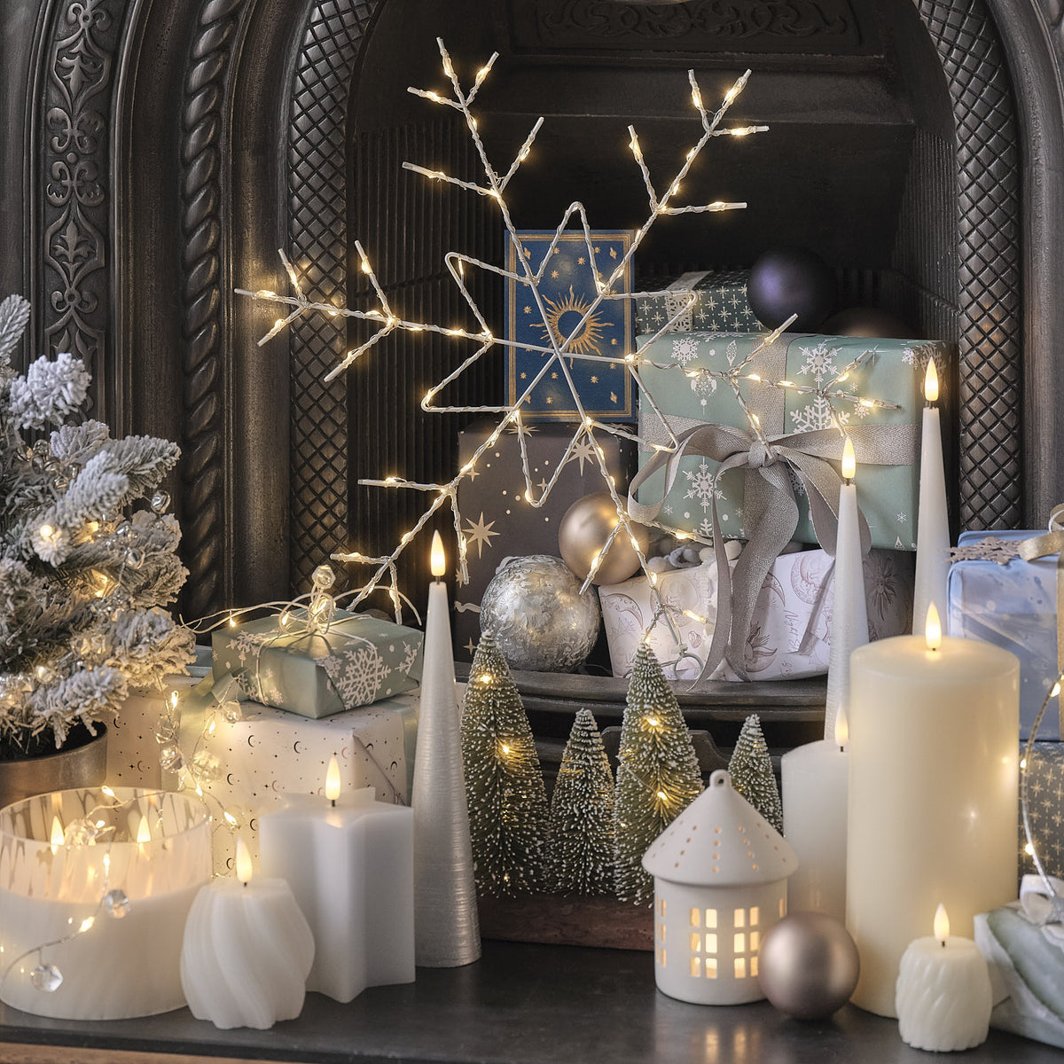 Schwarzer Kamineinsatz mit weißen LED-Kerzen, LED-Schneeflockenlicht-Motiv, grüne und silberne Weihnachtskugeln, silberner Rentier-Leuchtkörper, beleuchtete Häuschen-Ornamenten und weiße Leinenservietten.