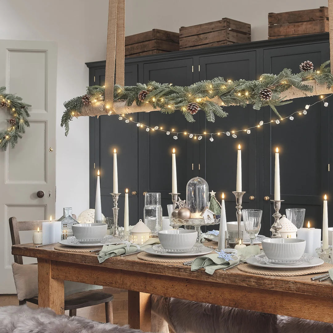 Festliche Weihnachtstischdekoration mit weißen LED-Kerzen in hoher Kegel-Form mit strukturierter Oberfläche, warmweißen LED-Flammen, weißen Taper-LED-Kerzen auf silbernem Kerzenständer, weißes Leinen, grüne Serviette, Weihnachtsgirlande, rote Beeren und Zapfen, Platzkartendekor auf Holztisch.