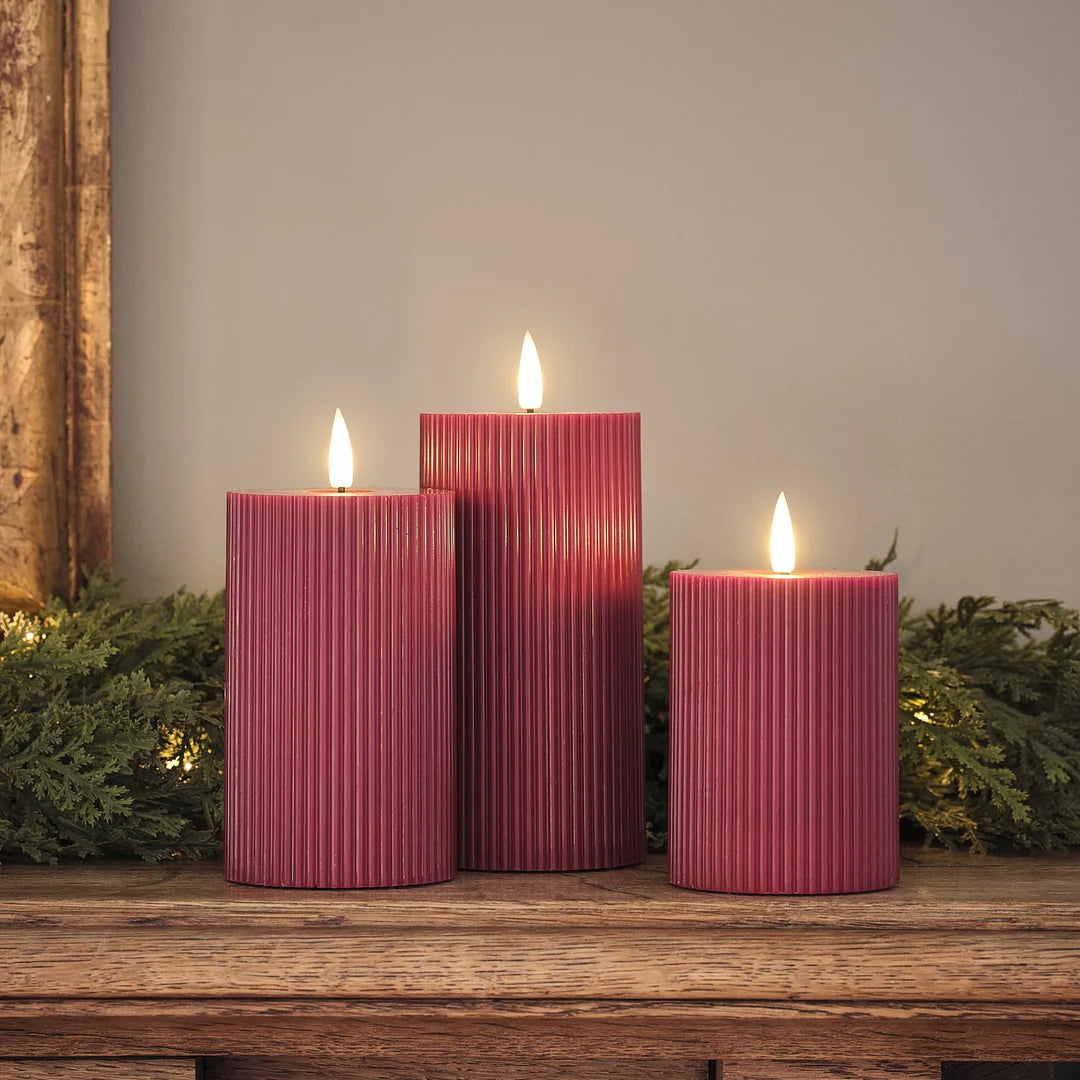 Drei rote LED-Zylinderkerzen in verschiedenen Höhen mit strukturierter Oberfläche und warmweißen LED-Flammen auf Holztisch, grüne Girlande mit goldenen Lichtern im Hintergrund.