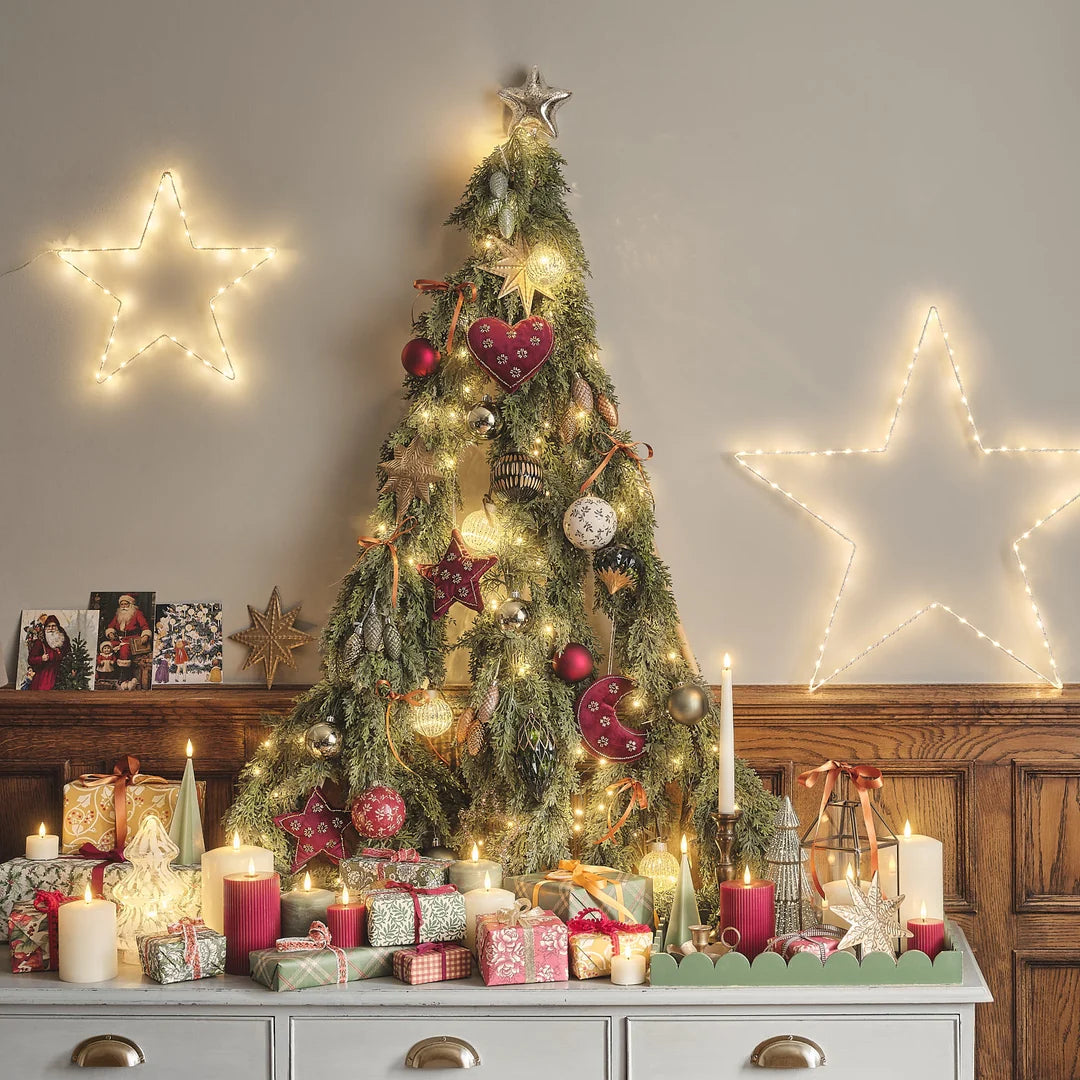 Weihnachtliches Tischdekor mit grünen LED-Taper-Kerzen, roten LED-Zylinderkerzen, weißer LED-Kerze mit dekorativem Muster, goldener Stern-Dekoration, Weihnachtskugeln in Burgunderrot und Weiß, Kerzenständer aus Messing, Girlande, Weihnachtsgeschenke, Holztisch.