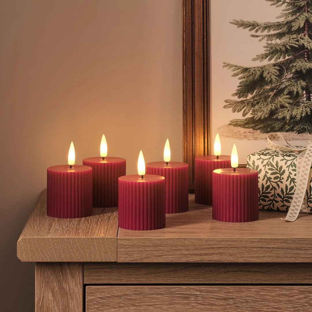 Sechs rote strukturierte LED-Zylinderkerzen mit warmweißen LED-Flammen auf gespaltener Holzoberfläche, beige und dunkles Holz, grüne Girlande mit goldenen Lichtern, dekoratives Leinen-Kissen mit Muster im Hintergrund.