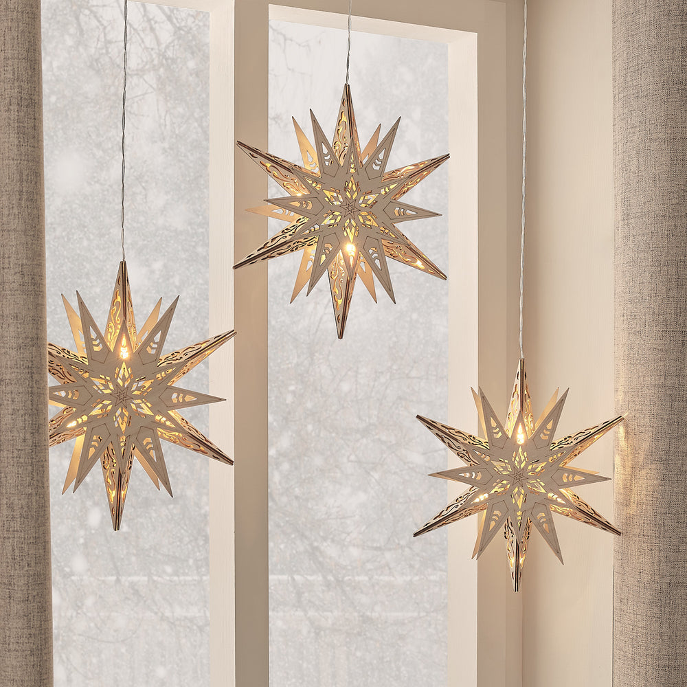 Drei warmweiß leuchtende LED-Holzsterne mit Schablonenmuster an dünnen Kabeln vor Fenster