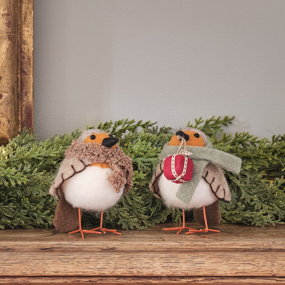 Zwei cremefarbene Vogel-Dekofiguren mit roten gestrickten Weihnachtsmützen und burgundy Schleife, stehend auf grünem Moosuntergrund mit Holzstamm-Hintergrund – niedliche Weihnachtsfiguren für Tischdekoration, Kaminsims und festliche Wohnzimmer-Arrangements mit warmem Charme.