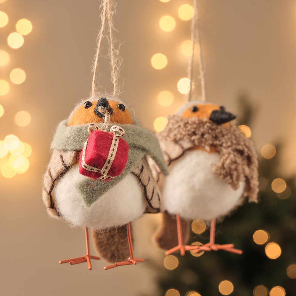 Drei cremefarbene hängende Vogel-Dekofiguren mit roten gestrickten Weihnachtsmützen, aufgehängt vor warmweißer LED-Lichterkette und weihnachtlichem Bokeh-Hintergrund – elegante Hängedekoration für Fenster, Weihnachtsbäume und festliche Indoor-Arrangements mit gemütlicher Beleuchtung.