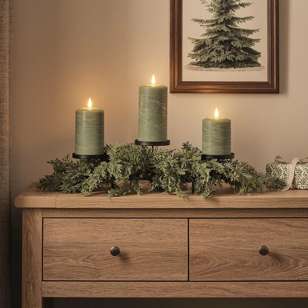Festliche Detailaufnahme eines hochwertigen Adventsgestecks mit schwarzen Kerzenhaltern und brennenden warmweißen LED-Kerzen, arrangiert auf Holzkommode mit dichtem grünem Fichtengrün, cremefarbenen Dekofiguren und gerahmtem Tannenbaum-Kunstdruck – luxuriöses Adventsarrangement mit authentischem Kerzenschein für stimmungsvolle elegante Weihnachtsdekoration.