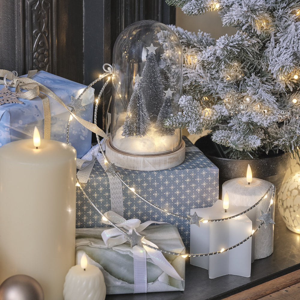 LED-Glasglocke mit Weihnachtsbäumen auf Geschenkpaketen neben Kerzen und Lichterkette mit Sternen