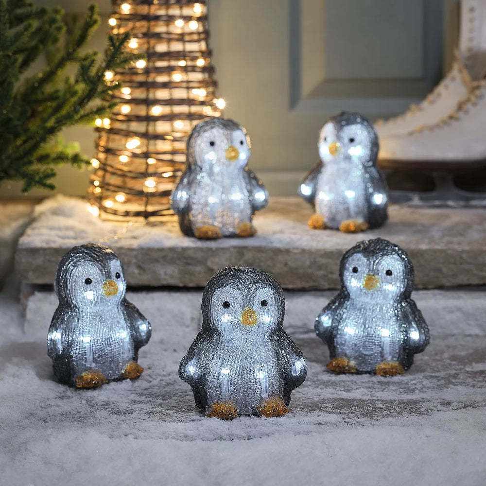 Gruppe von glitzernden grau-weißen Pinguin-Dekofiguren mit Schneeeffekt-Oberflächenfinish und goldenen Details, arrangiert auf grauem Stein-Untergrund vor unscharfem grünem Hintergrund – liebevolle Pinguin-Familie für klassische Weihnachtsarrangements und festliche Außendekoration.