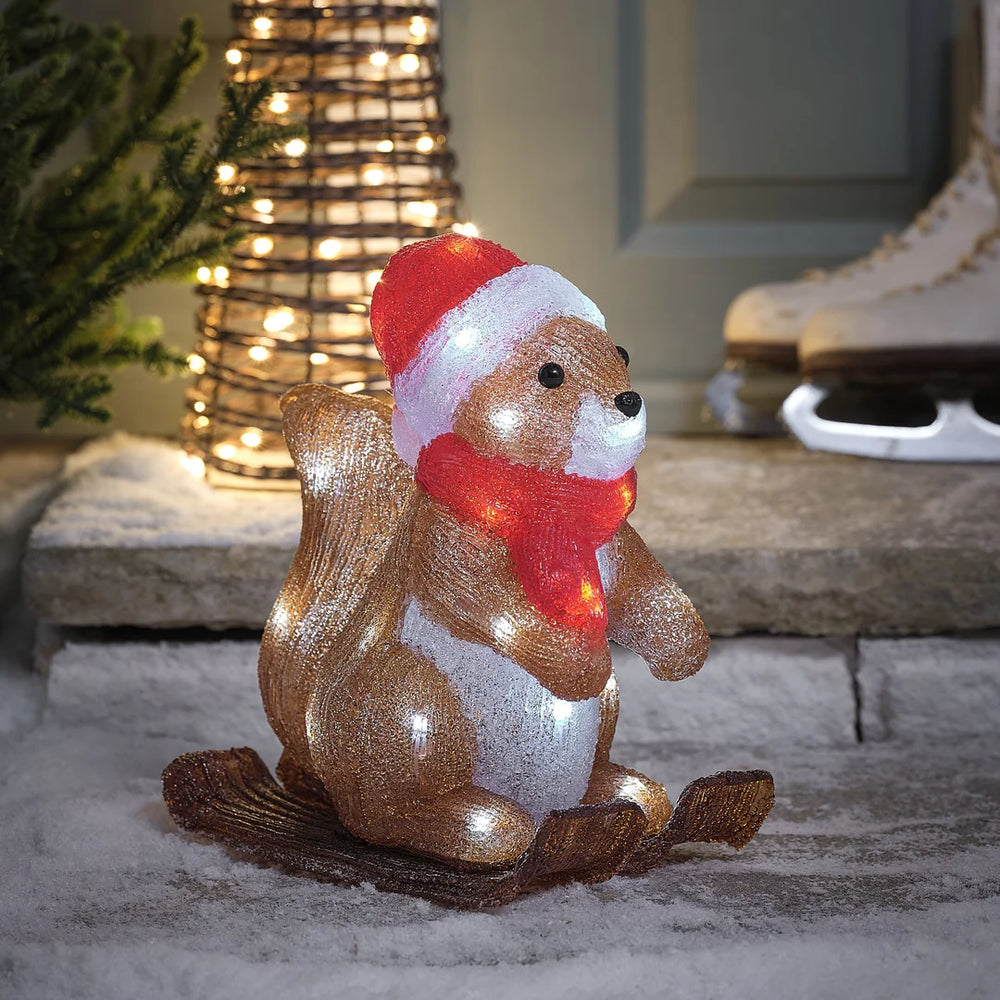Charmante glitzernde braun-goldene Eichhörnchen-Dekofigur mit roter gestrickter Weihnachtsmütze und rotem Schal, positioniert vor beleuchteter Kegel-Weihnachtsbaumdekoration auf Stein-Terrasse – festliche Waldtier-Figur für Außendekoration, Garten-Arrangements und weihnachtliche Hausdekorationen mit eleganter Glitter-Textur und warmweißer LED-Beleuchtung im Hintergrund.