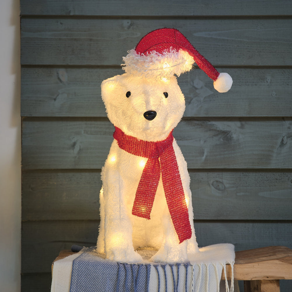 Charmante cremefarbene Plüsch-Teddybär-Figur mit roter gestrickter Weihnachtsmütze und rotem Textilschal, positioniert auf blauem Holzuntergrund vor dunklem Holzwand-Hintergrund – festliche Plüsch-Dekofigur für Außendekoration, Garten-Arrangements und weihnachtliche Hausdekorationen mit klassischem Weihnachtsstil.