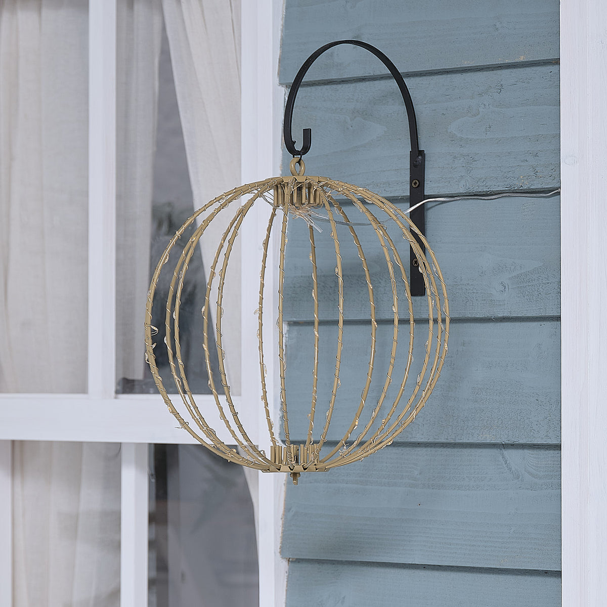 Elegante hängende LED-Kugel-Dekofigur mit offener Drahtstruktur in geometrischem Design, positioniert an schwarzem Metallbogen vor hellblauem Holzwand-Hintergrund – dekorative Hängeform für Außendekoration und klassische Weihnachtsarrangements mit modernem Drahtdesign.