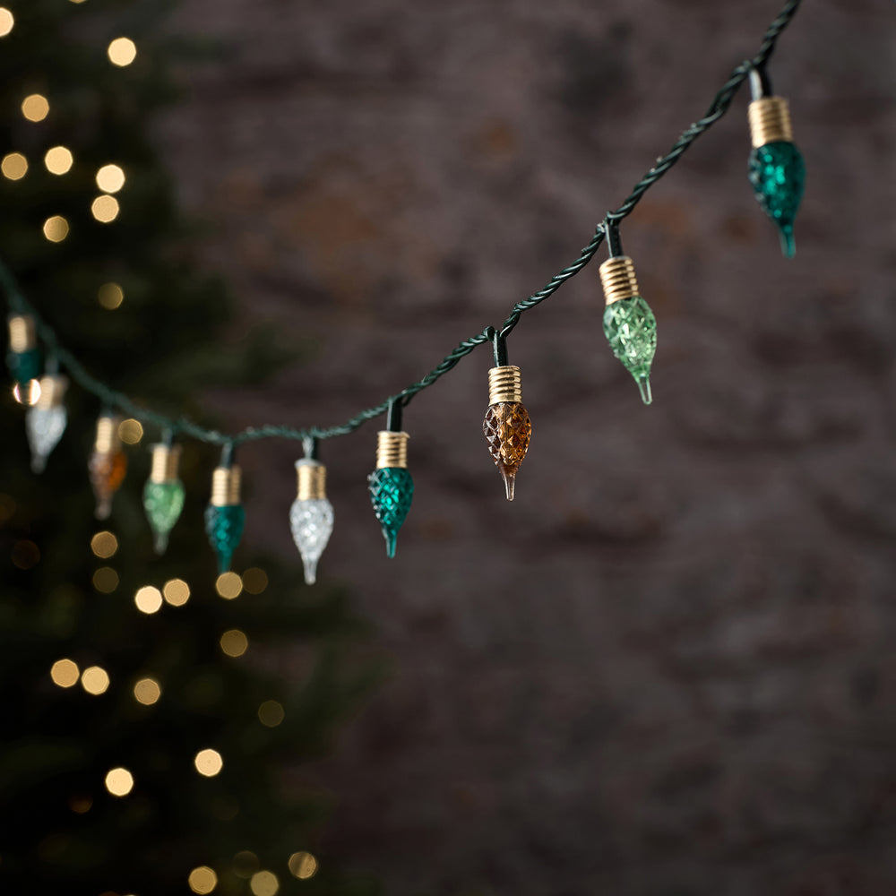Premium-LED-Lichterkette mit schwarzem Draht und hängenden Tropfen-Anhängern in Regenbogenfarben (Grün, Gold, Orange, Weiß), arrangiert vor unscharfem Bokeh-Lichthintergrund – dekorative mehrfarbige Lichterkette für elegante Außen-Weihnachtsarrangements und gehobene Fassaden-Dekorationen.