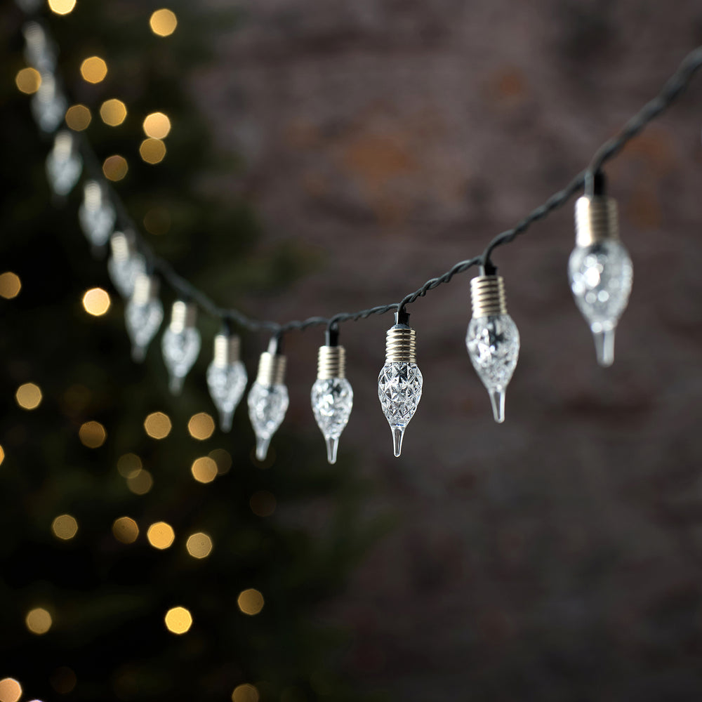Premium-LED-Lichterkette mit schwarzem Draht und hängenden kristallinen Tropfen-Anhängern in eleganter Champagner-Gold-Färbung, arrangiert vor unscharfem Bokeh-Lichthintergrund – luxuriöse Lichterkette für elegante Außen-Weihnachtsarrangements und gehobene Fassaden-Dekorationen.