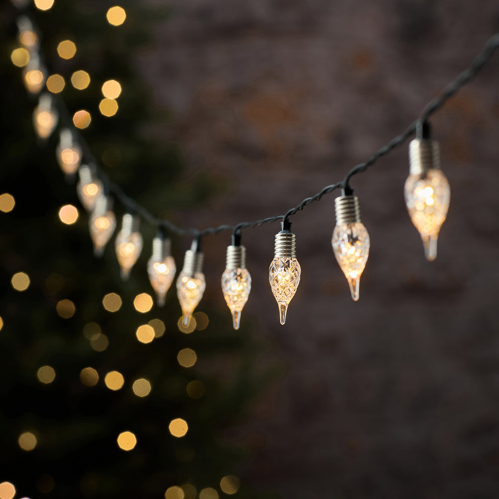 Elegante LED-Lichterkette mit schwarzem Draht und hängenden kristallinen Tropfen-Dekofiguren in Champagner-Gold-Tönen, mit warmweißem Bokeh-Hintergrund und unscharfen Lichtern – festliche Premium-Lichterkette für Außendekoration, Veranda-Arrangements und weihnachtliche Hausdekorationen mit luxuriösem Glühen.