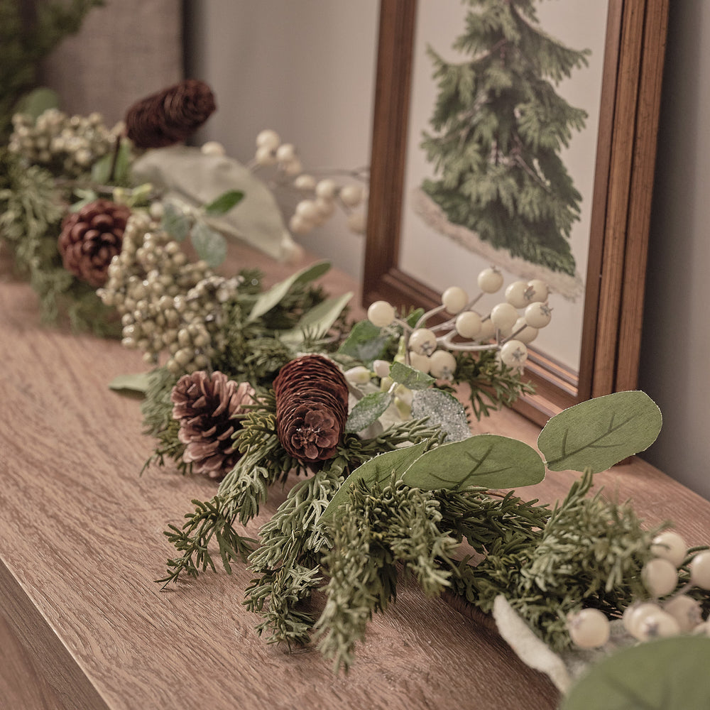 Charmantes üppiges Fensterbrett-Arrangement mit grünem Fichtengrün, burgundy strukturierten Blüten, weißen Beeren und natürlichen Holzakzenten, positioniert auf hellbraunem Holztisch vor Fensterrahmen mit Tannenbaum-Kunstdruck – festliche Weihnachts-Tischdekoration für Fensterdekorationen und elegante Wohnzimmer-Displays mit natürlichem Waldcharme.