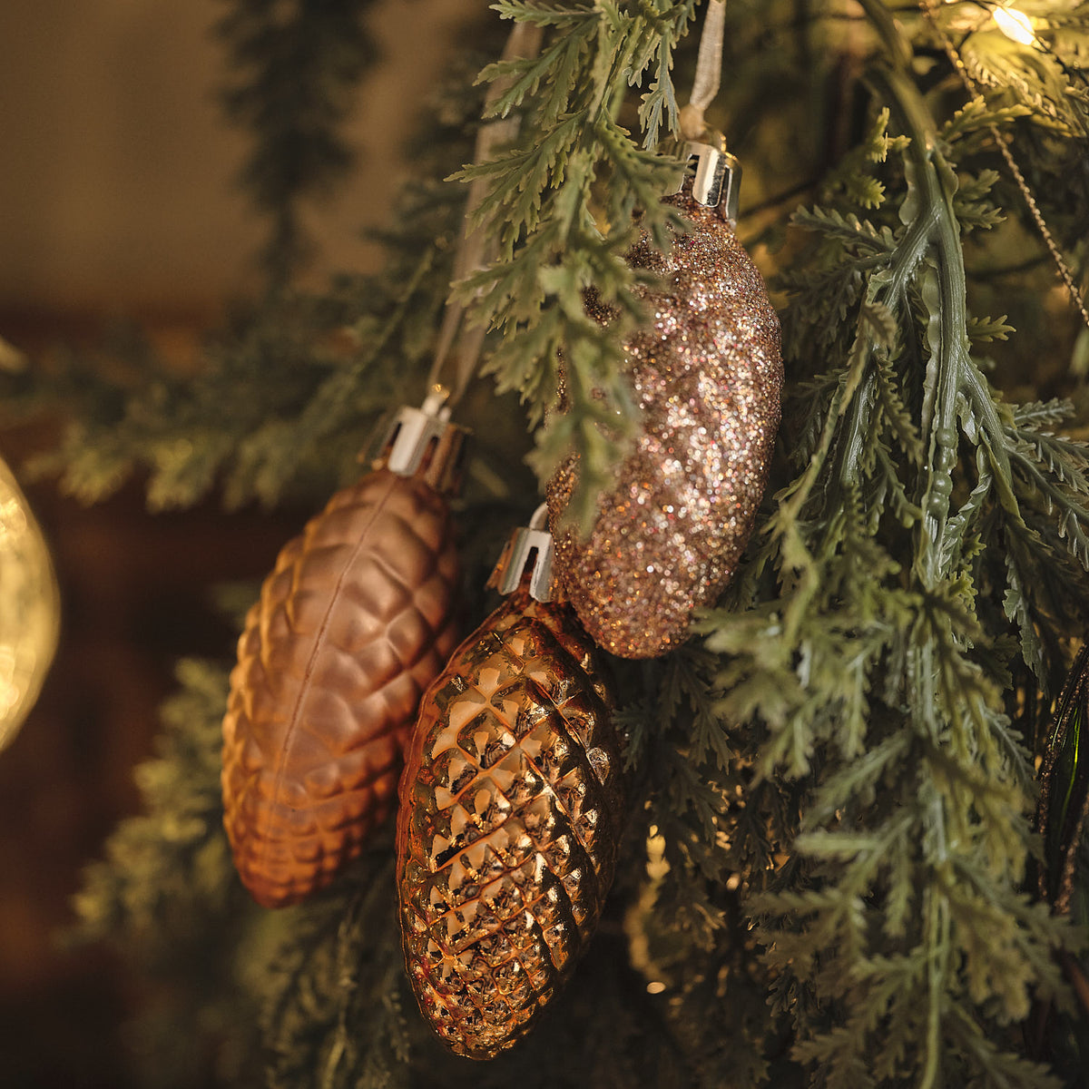 Natürliche Tannenzapfen in Braun- und Kupfertönen an grünen Zweigen mit warmer Beleuchtung, Detailansicht von hängenden Weihnachtsdeko-Elementen.
