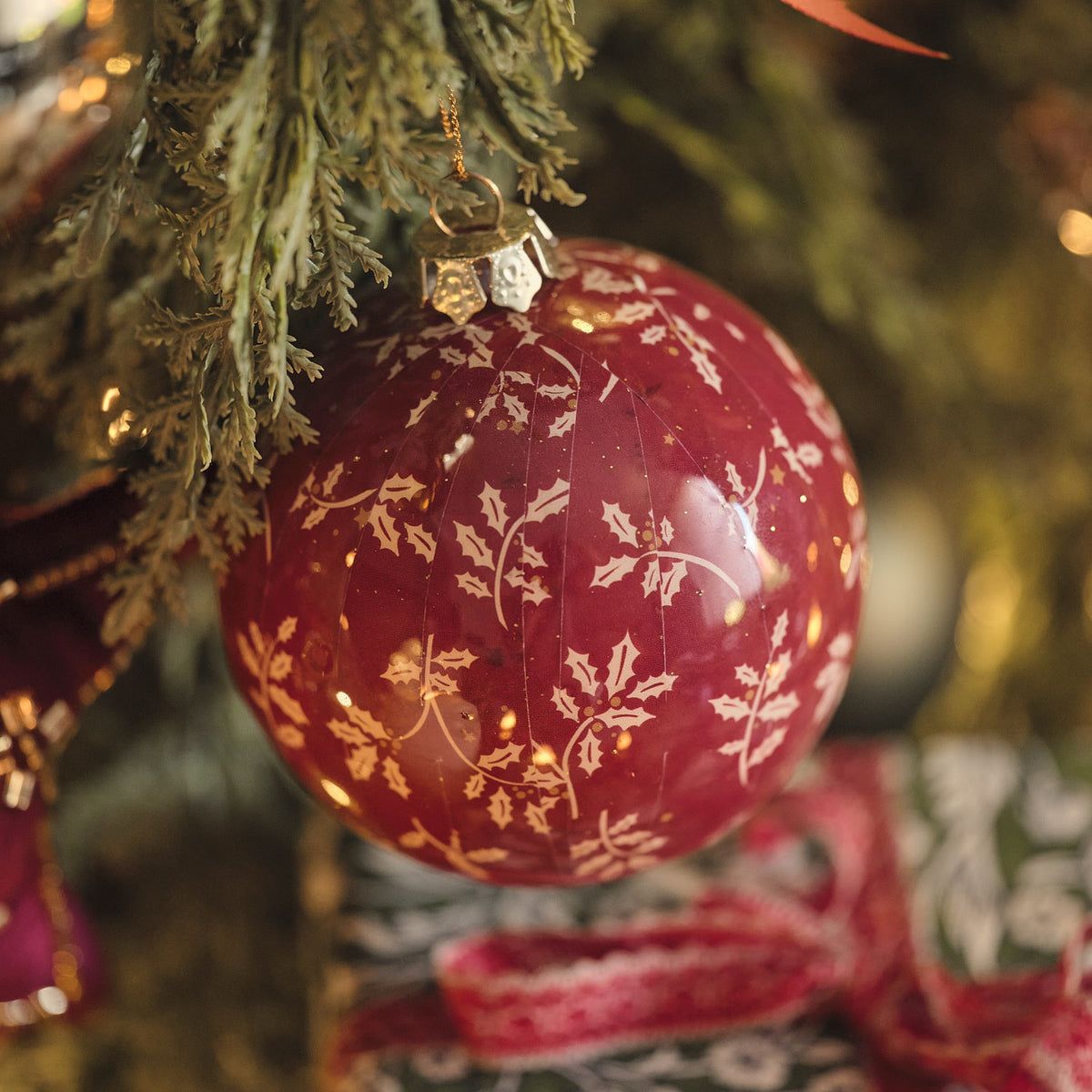 Bordeauxrote Weihnachtskugel mit weißem Blumenmuster an Tannenzweig mit roter Schleife.
