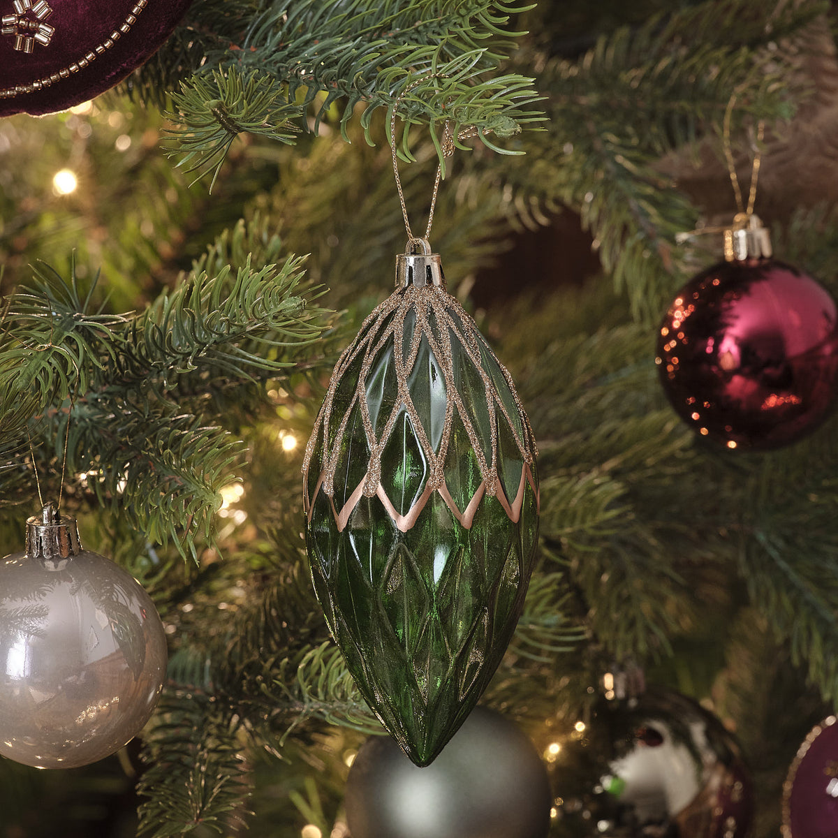 Grün-goldene Weihnachtskugel in Tropfenform mit Rautenmuster am Tannenzweig neben roten und weißen Kugeln.