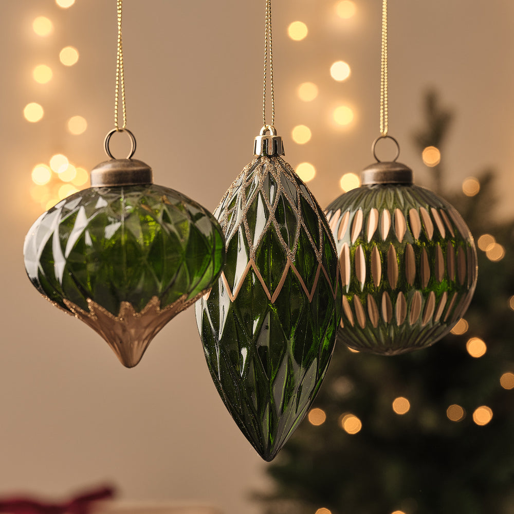 Grün-goldene Glas-Weihnachtskugeln in Tropfen- und Rautenform mit facettiertem Muster, hängend vor Lichterglanz.