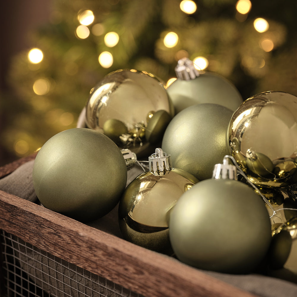 Detailansicht olivgrüner und goldener Weihnachtskugeln in matter und glänzender Optik in Holzkiste vor Lichterglanz.