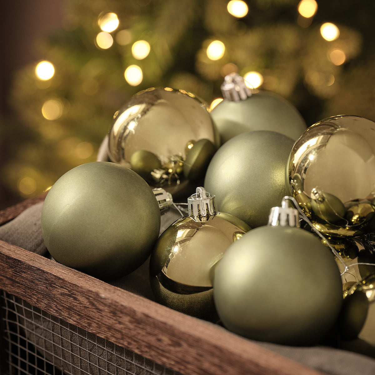 Detailansicht olivgrüner und goldener Weihnachtskugeln in matter und glänzender Optik in Holzkiste vor Lichterglanz.