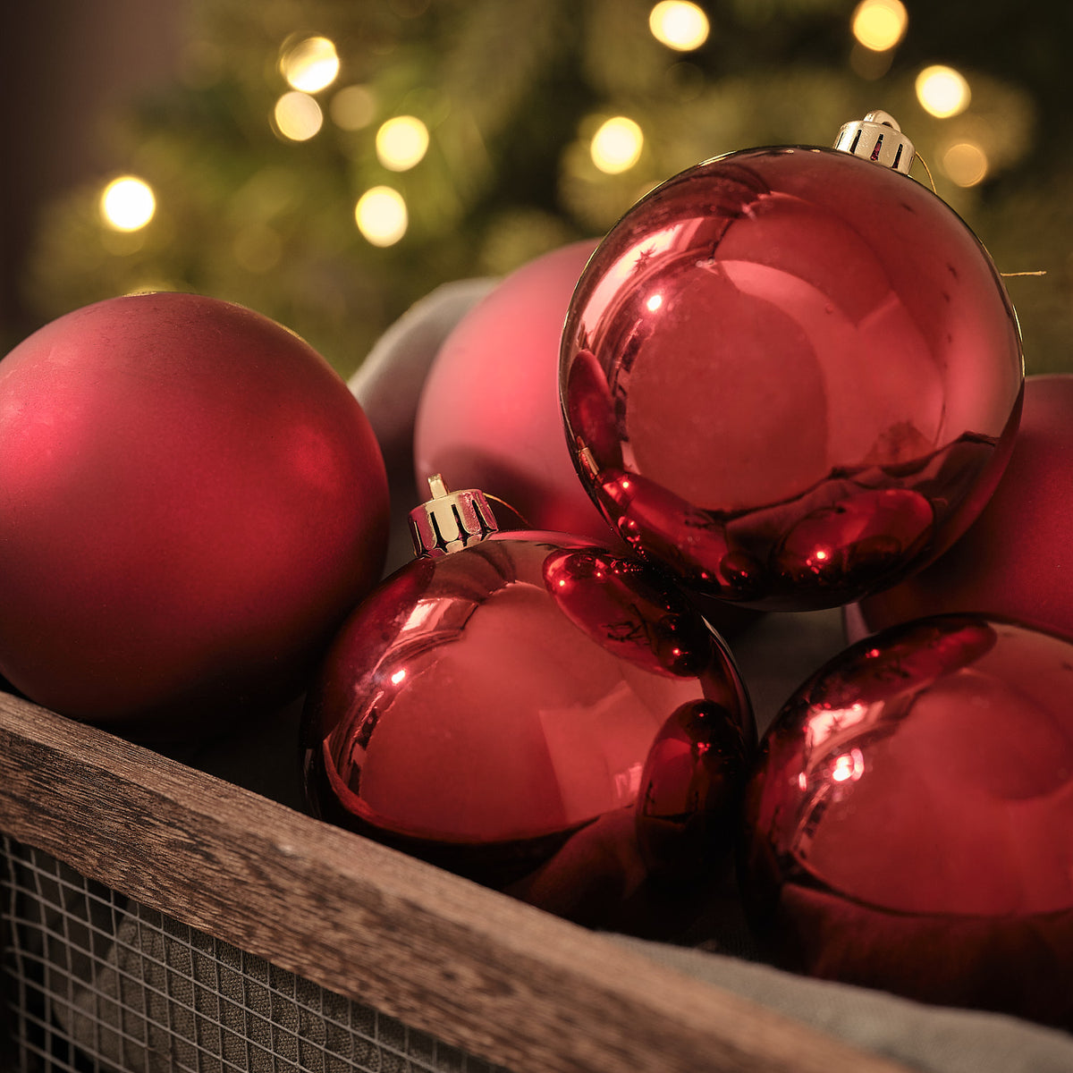 Glänzende rote Weihnachtskugeln in Holzkiste vor beleuchtetem Tannenbaum.