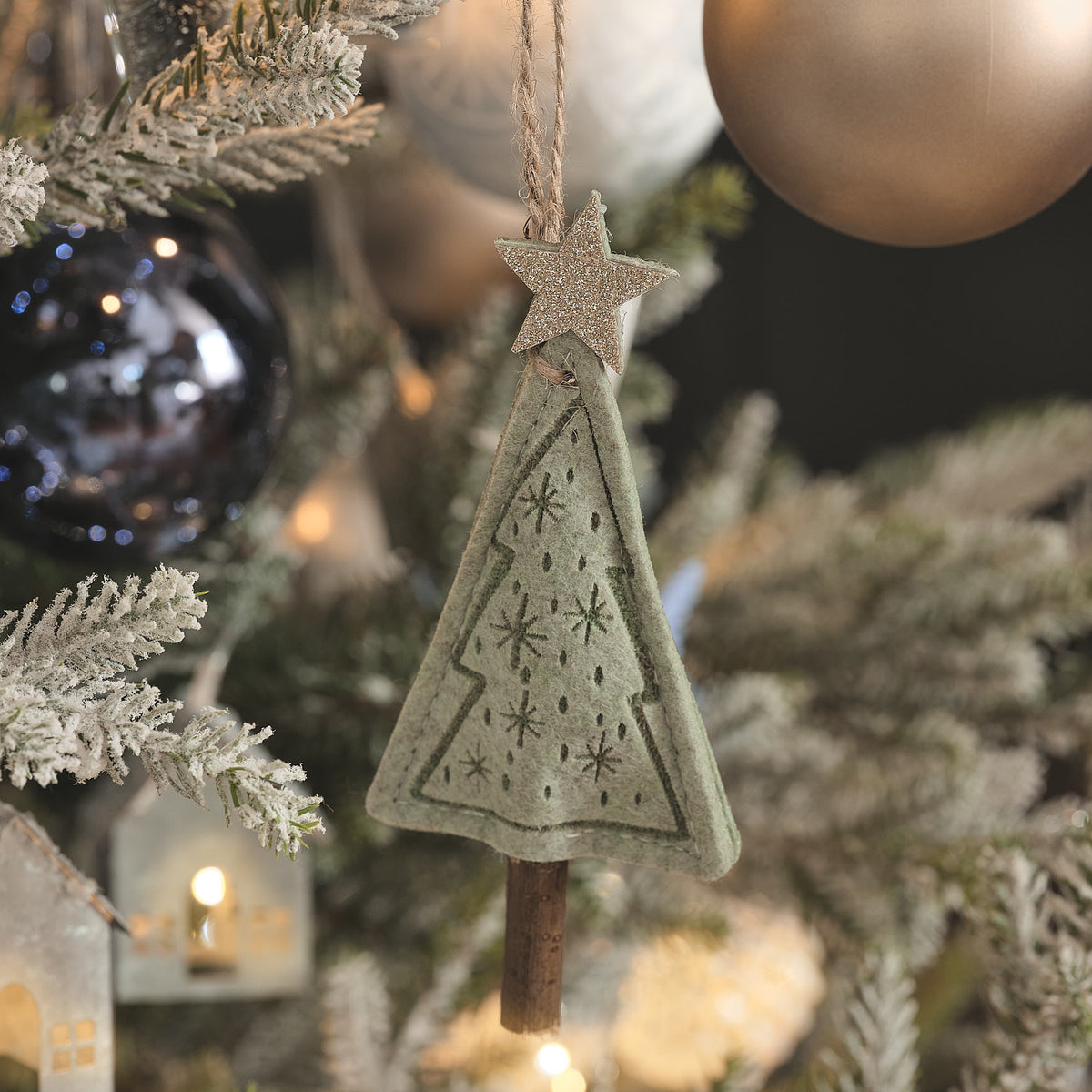 Grüner Stoff-Tannenbaum-Anhänger mit Sternspitze an bereiftem Tannenzweig neben dunkler Kugel.