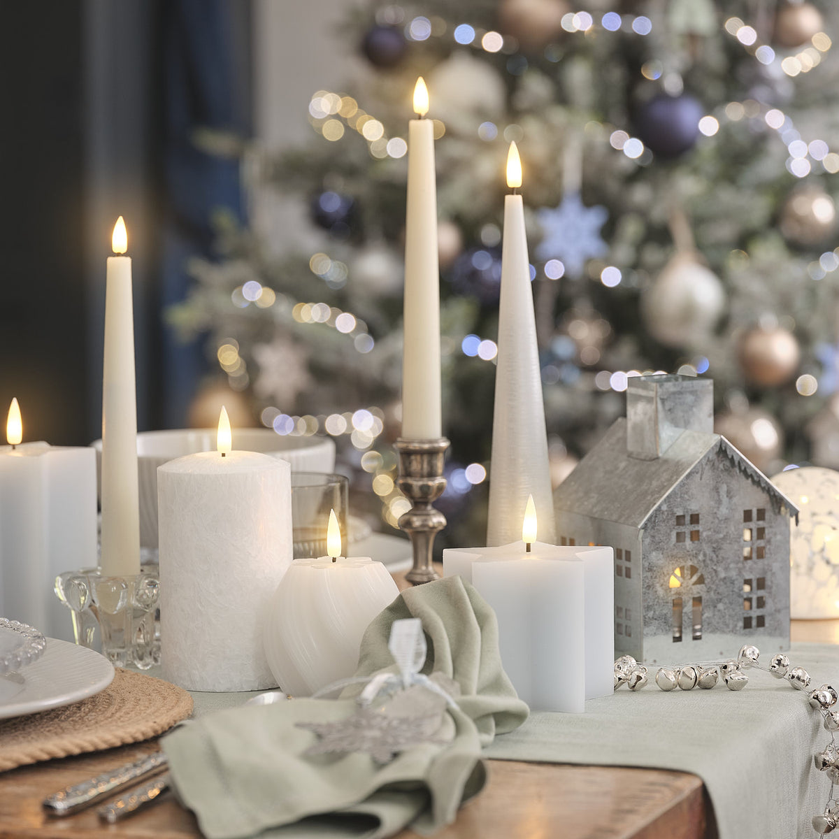 Perlenlichterkette als Tischdekoration mit weißen Kerzen und Weihnachtsbaum im Hintergrund.