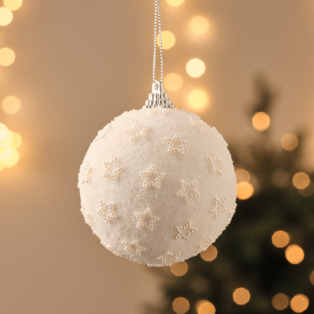Weiße Weihnachtskugel mit Perlen- und Sternenbesatz, hängend vor beleuchtetem Tannenbaum.