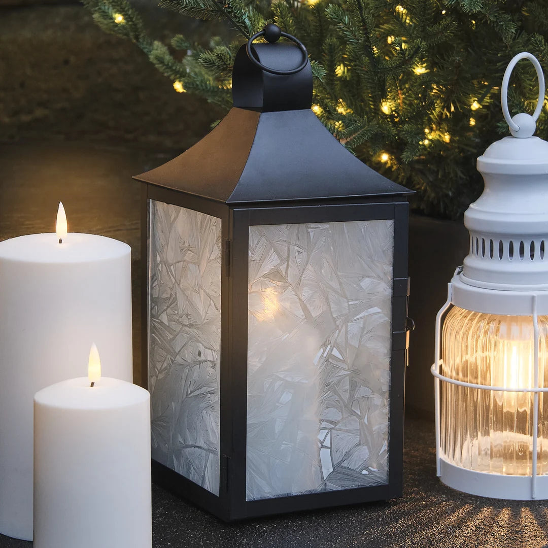 Schwarze LED-Laterne neben weißen Kerzen und weiterer Laterne vor Weihnachtsbeleuchtung.