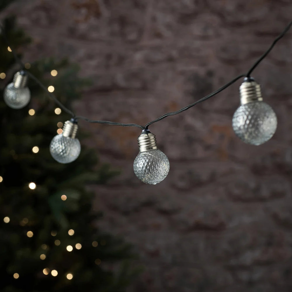 LED-Lichterkette mit warmweißen und silbernen strukturierten Glühbirnen vor Tannenzweig.