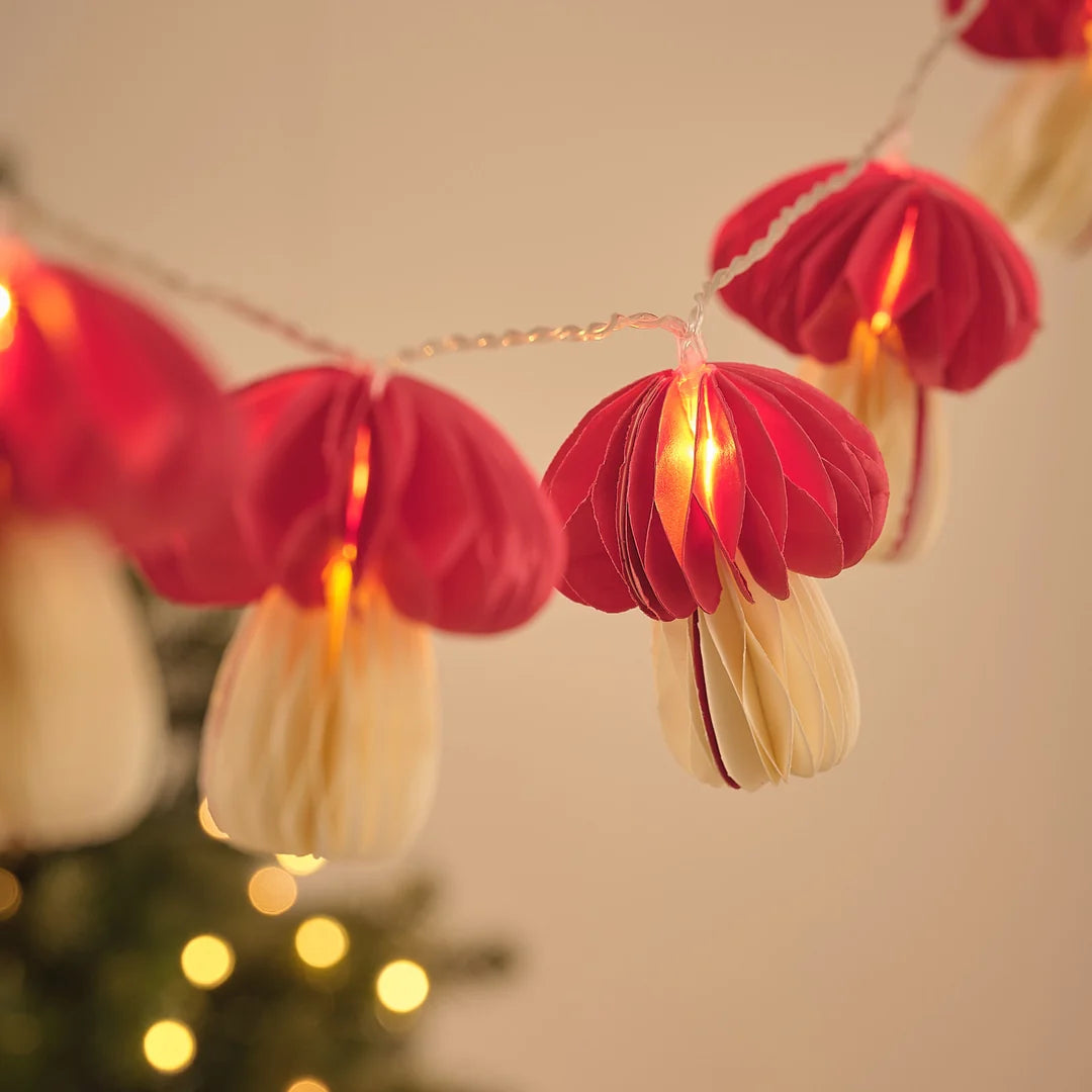 Lichterkette mit roten Papier-Pilz-Anhängern vor Weihnachtsbaum und Lichterglanz.