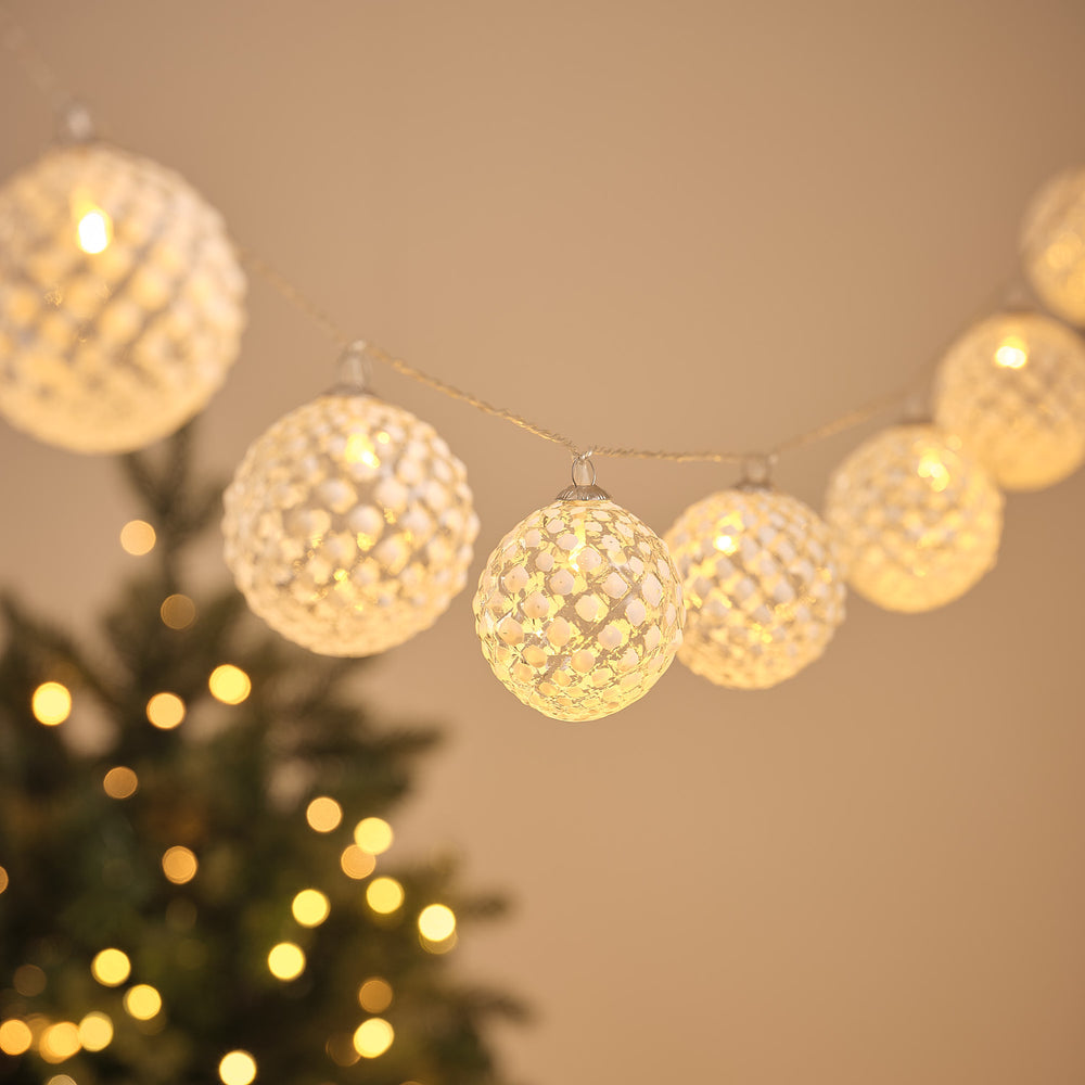 LED-Lichterkette mit zehn warmweiß leuchtenden, gefrosteten Glaskugeln vor unscharfem Weihnachtsbaum