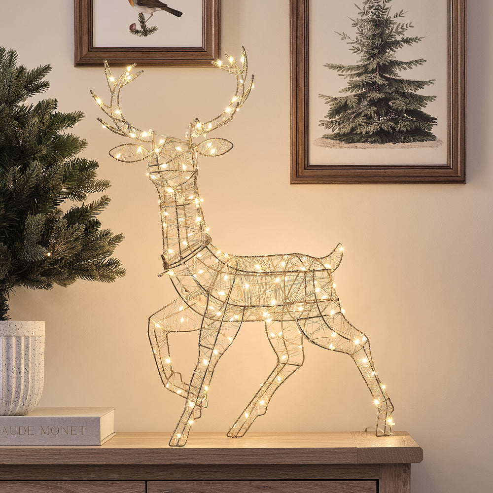 Goldener LED-Hirsch Hutton Mini mit warmweiß leuchtenden Mikro-LEDs auf Holzkommode vor Wand mit Bildern