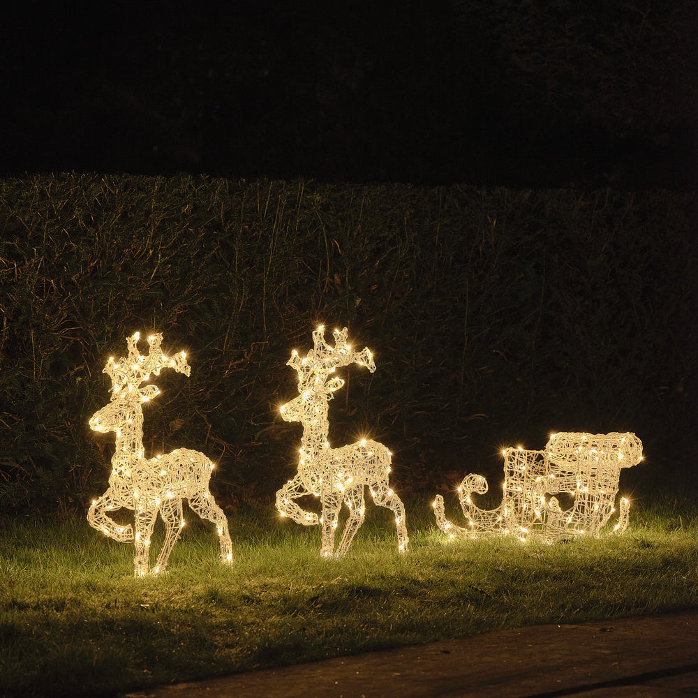 Zwei warmweiß beleuchtete LED-Rentiere mit Schlitten auf Rasen vor dunkler Hecke bei Nacht