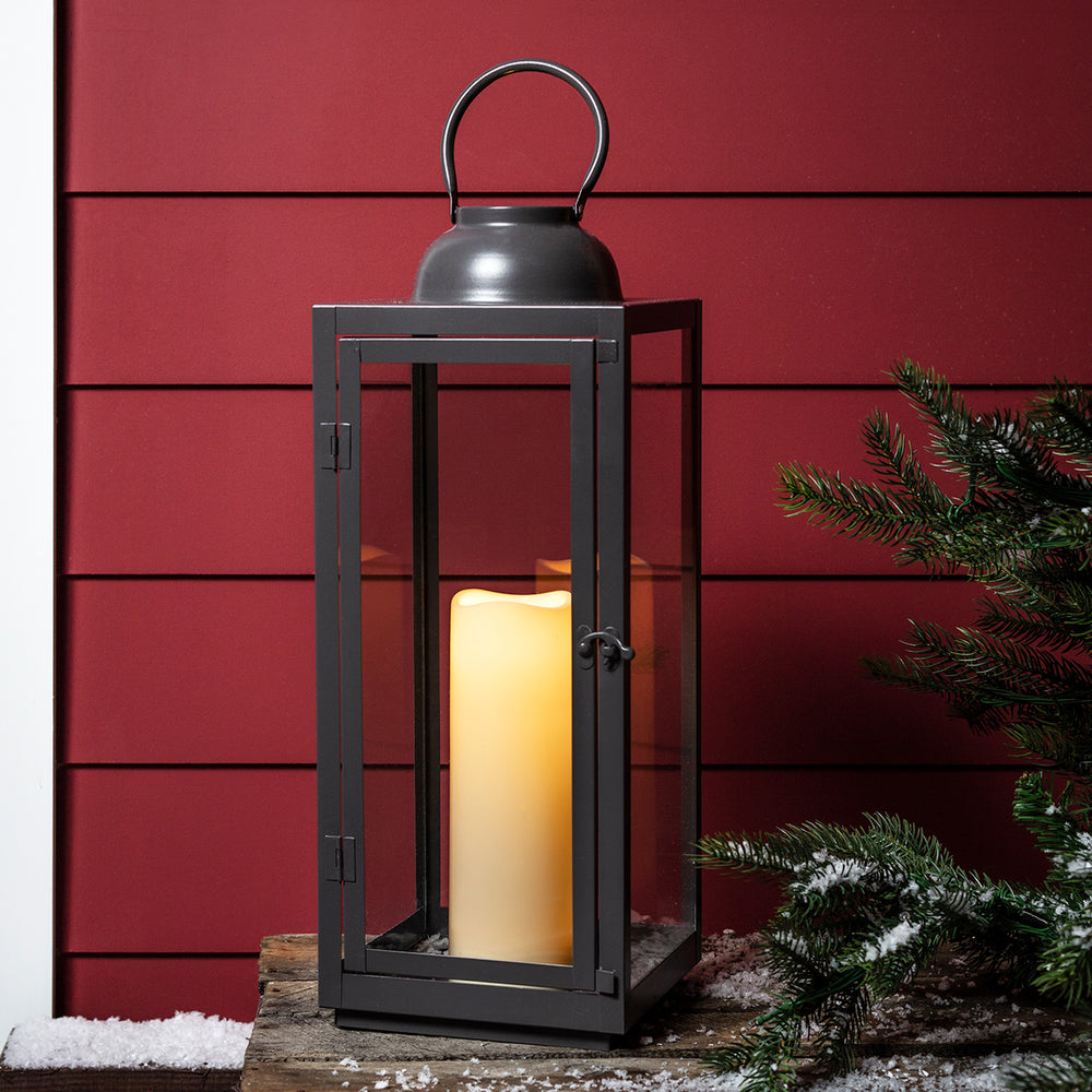 Hohe graue Gartenlaterne mit LED-Kerze vor roter Hauswand.
